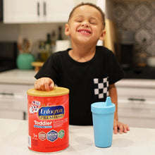 Child holding a can of Enfagrow Premium Toddler Nutritional Milk Powder with a blue sippy cup on a kitchen counter.